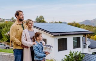 Två vuxna och ett barn står framför ett hus med solceller. Barnet håller i en pappersmodell av ett hus med solceller.