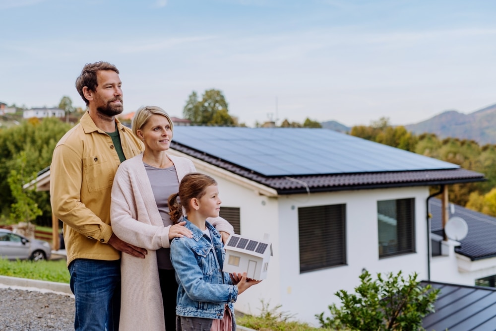 Två vuxna och ett barn står framför ett hus med solceller. Barnet håller i en pappersmodell av ett hus med solceller.